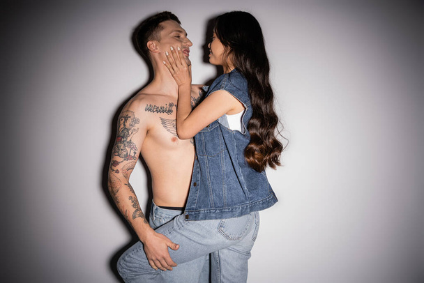 smiling asian woman in denim clothes touching face of tattooed muscular man on grey background - Foto, immagini