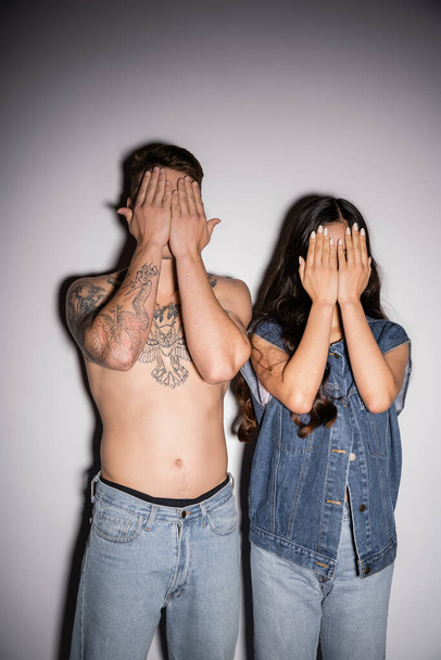 brunette woman in denim vest and shirtless man with tattooed body obscuring faces with hands on grey background - 写真・画像