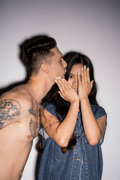 tattooed man kissing brunette asian woman obscuring face with hands on grey background - Photo, Image