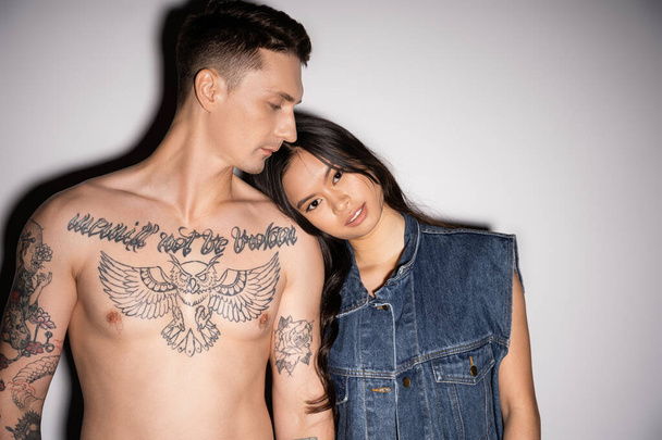 asian woman in denim vest looking at camera while leaning on shirtless tattooed man on grey background - Foto, immagini