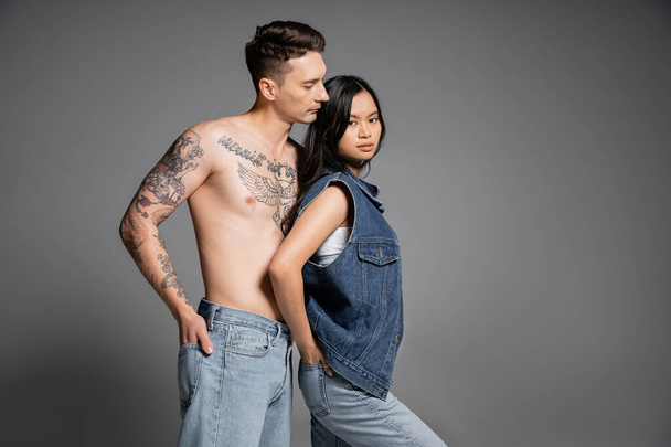 morena asiática mulher e tatuado sem camisa homem de pé com as mãos em bolsos traseiros de jeans no fundo cinza - Foto, Imagem