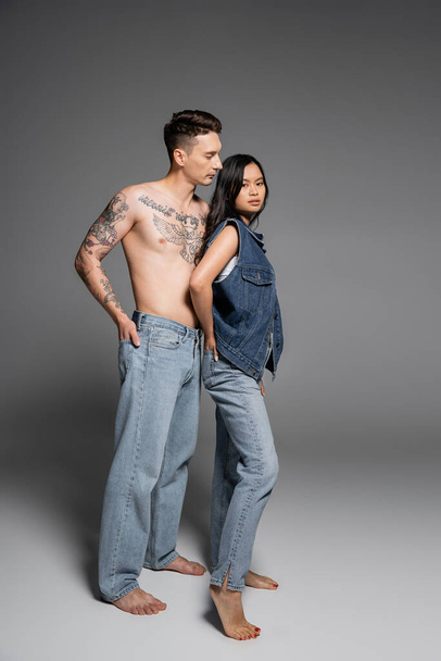 full length of barefoot tattooed man in jeans near brunette asian woman in denim outfit posing on grey background - Photo, Image