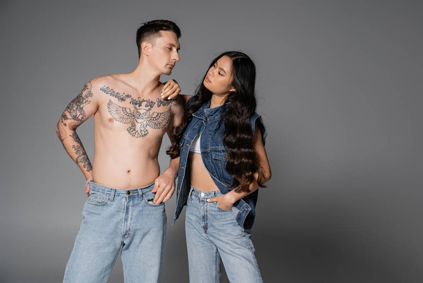 asian woman in stylish denim outfit looking at shirtless tattooed man in jeans on grey background - Photo, image