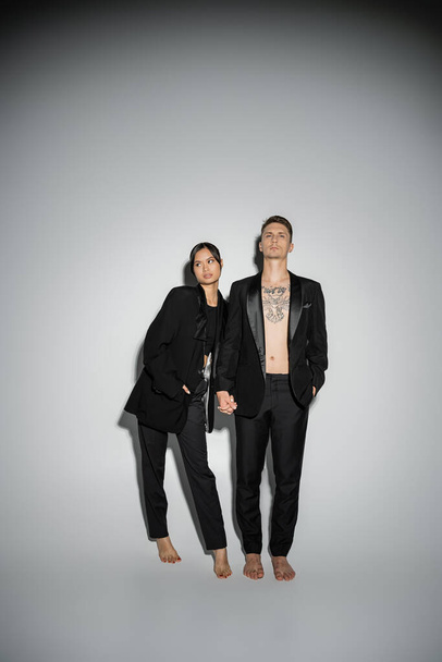 full length of brunette asian woman and tattooed man posing in black stylish suits on grey background - Foto, afbeelding
