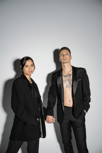 stylish interracial couple in black elegant blazers holding hands and looking at camera on grey background - Foto, afbeelding