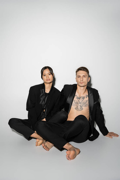 full length of barefoot interracial couple in black elegant pantsuits sitting with crossed legs on grey background - Fotó, kép