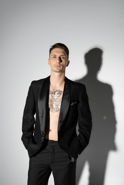 front view of shirtless tattooed man in black elegant pantsuit posing with hands in pockets on grey background with shadow - Fotó, kép