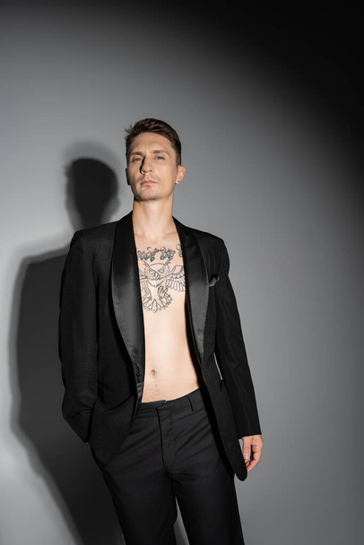shirtless tattooed man in black blazer and pants looking at camera on grey background with shadow - Photo, Image
