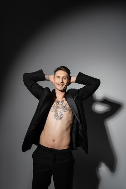 Ευτυχισμένος άντρας με μαύρο σακάκι σε σώμα χωρίς πουκάμισο με τατουάζ κρατώντας τα χέρια πίσω από το κεφάλι σε γκρι φόντο με σκιά - Φωτογραφία, εικόνα