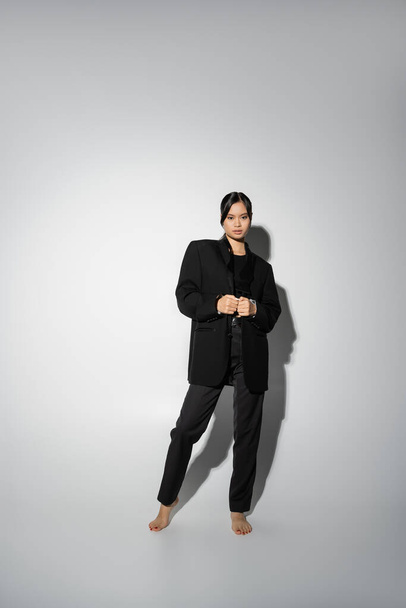 comprimento total de mulher asiática descalça vestindo terno elegante preto enquanto olha para a câmera no fundo cinza com sombra - Foto, Imagem
