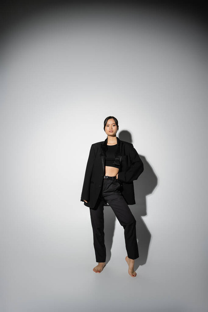 comprimento total de descalço mulher asiática em preto elegante traje posando com a mão na cintura no fundo cinza com sombra - Foto, Imagem