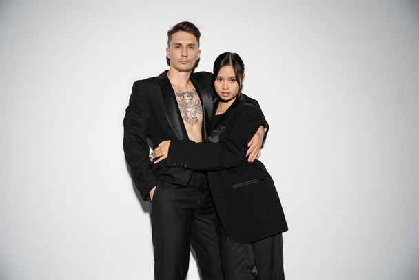 moda casal multiétnico em ternos elegantes pretos abraçando e olhando para a câmera no fundo cinza - Foto, Imagem