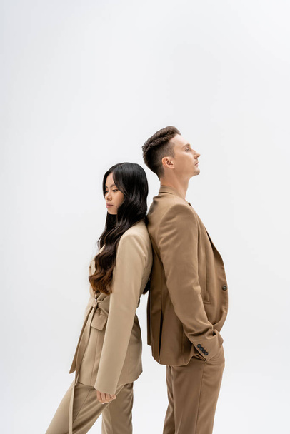 side view of interracial couple in beige suits standing back to back isolated on grey - 写真・画像