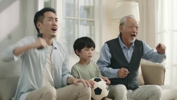 Ασιάτης γιος πατέρας και παππούς βλέποντας ζωντανά μετάδοση ποδοσφαιρικού αγώνα στην τηλεόραση μαζί στο σπίτι και απογοητευμένος από την απόδοση της ομάδας τους - Πλάνα, βίντεο
