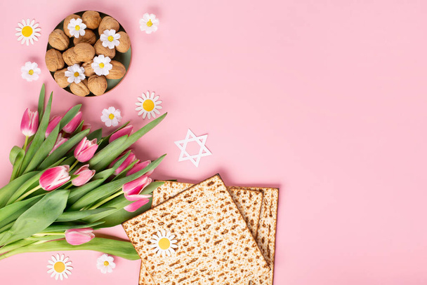 ユダヤ人の休日ピンクのテーブルの上にマツア、ナッツ、チューリップやデイジーの花と過越のグリーティングカードの概念。Seder Pesach春休みの背景、トップビュー、コピースペース. - 写真・画像