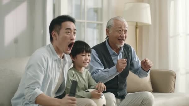 asian son father and grandfather watching live broadcasting of football match on TV together at home celebrating a goal - Filmati, video
