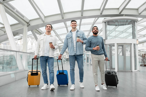 Διακοπές, επιτέλους. Τρεις Φίλοι Άνδρες Ταξιδεύοντας στο εξωτερικό Περπάτημα με βαλίτσες και κρατώντας χάρτινα φλιτζάνια καφέ Posing στο σύγχρονο αεροδρόμιο Terminal Indoor. Τουριστική ιδέα. Πλήρες μήκος - Φωτογραφία, εικόνα