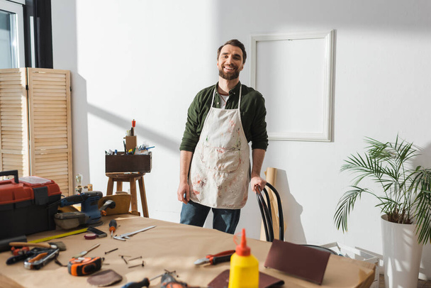 Smiling craftsman in apron standing near blurred tools on table  - Foto, immagini