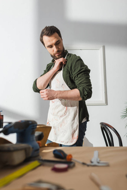 Bearded craftsman wearing apron near blurred tools on table in workshop  - Zdjęcie, obraz
