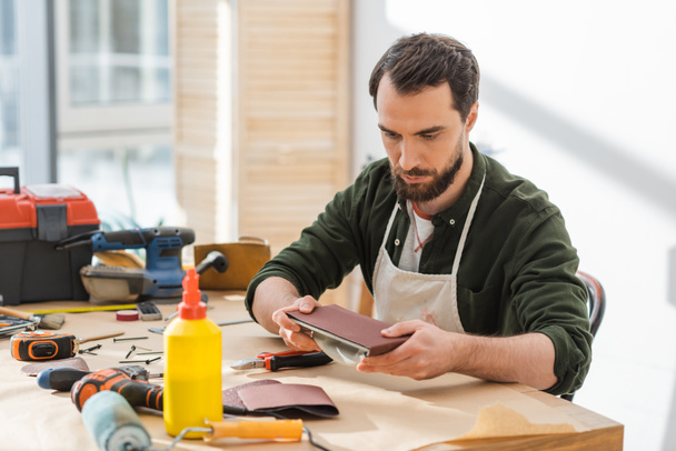 Carpintero barbudo en delantal sosteniendo papel de lija cerca de herramientas en la mesa en el taller  - Foto, imagen