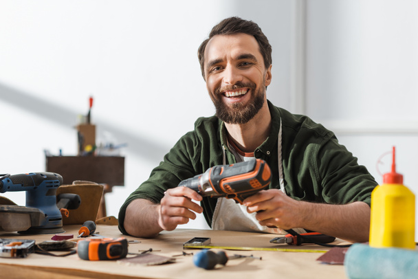 Веселый плотник держит электрическую отвертку и смотрит в камеру в мастерской  - Фото, изображение