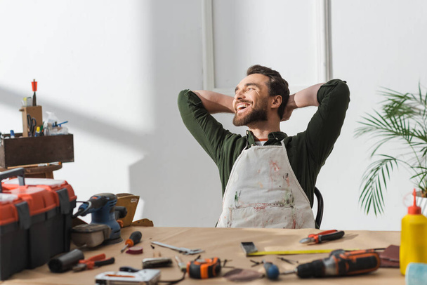 Cheerful craftsman in dirty apron resting near blurred tools on table in workshop  - Zdjęcie, obraz