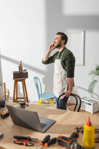 Positive craftsman talking on cellphone near tools and laptop on working table  - 写真・画像