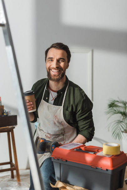 Lächelnder Tischler mit Kaffee zum Werkzeugkasten in der Werkstatt  - Foto, Bild