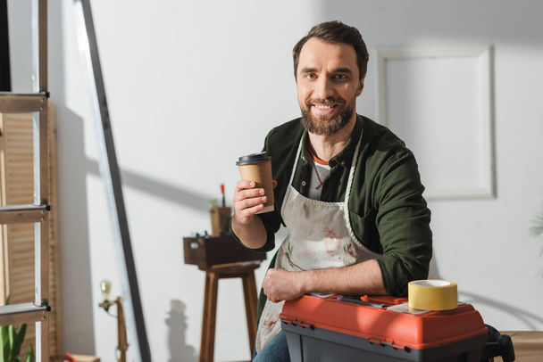 Cheerful carpenter in apron holding takeaway coffee near toolbox in workshop  - Fotoğraf, Görsel