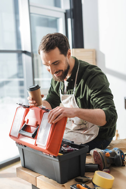 Önlüklü mutlu marangoz elinde kahve ve atölyedeki alet kutusunu açıyor.  - Fotoğraf, Görsel