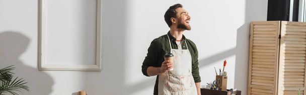 Smiling bearded repairman in apron holding paper cup in workshop, banner  - Foto, imagen