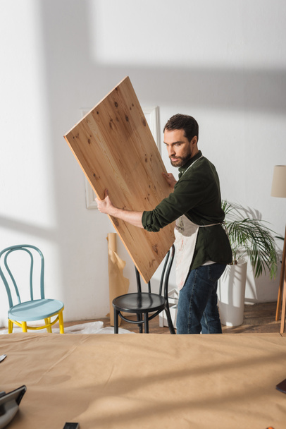 Плотник держит деревянную доску возле стульев в мастерской  - Фото, изображение