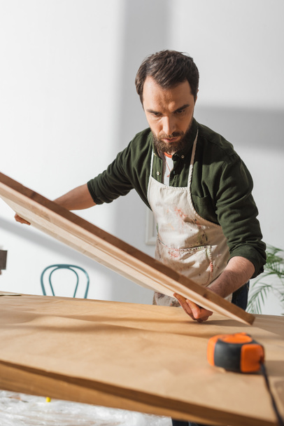 Artisan barbu dans un tablier tenant une planche de bois près de la règle dans l'atelier  - Photo, image