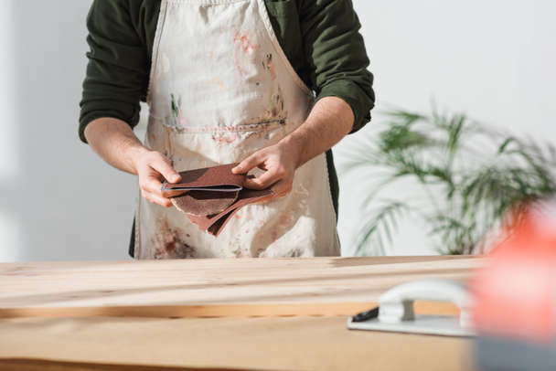 Обрезанный вид рабочего в фартуке с наждачной бумагой рядом с деревянной доской  - Фото, изображение