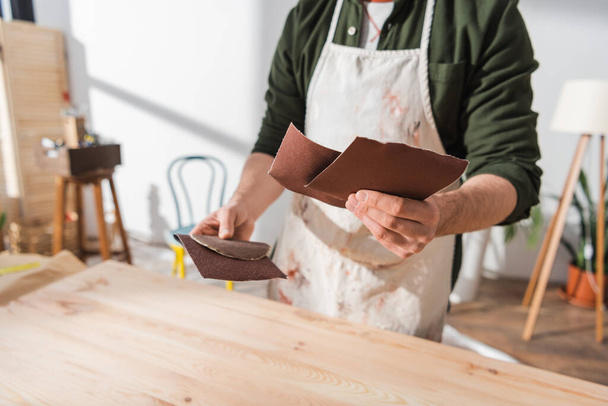 Vista recortada del artesano borroso en delantal sosteniendo papel de lija mientras trabaja con tablero de madera  - Foto, Imagen