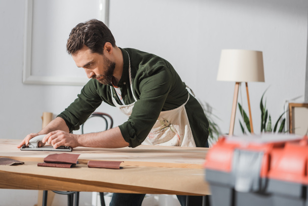 Reparador en delantal lijado tablón de madera cerca de la caja de herramientas borrosa en el taller  - Foto, Imagen