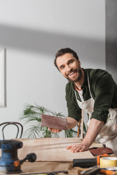 Carpintero sonriente en delantal sosteniendo papel de lija cerca de la tabla y mirando a la cámara en el taller  - Foto, imagen
