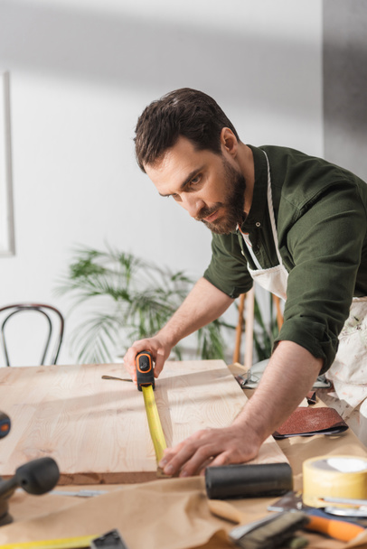 Restaurateur en tablier mesurant planche en bois avec règle près des outils en atelier  - Photo, image