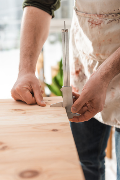 Vista ritagliata di artigiano in grembiule sporco misura tavola di legno con pinze in officina - Foto, immagini