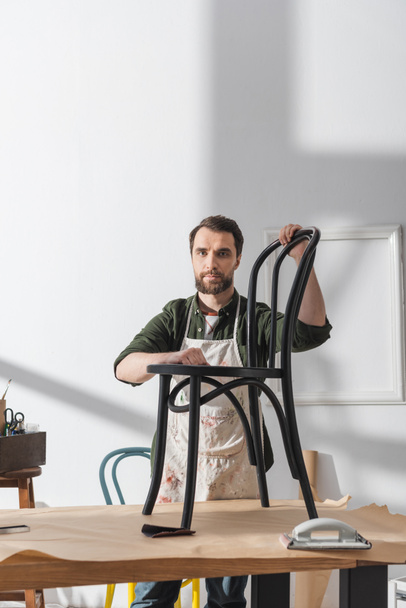 Бородатый реставратор в фартуке держит стул и смотрит на камеру рядом с рабочим столом в мастерской  - Фото, изображение