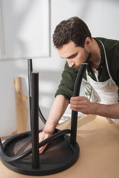 τεχνίτης σε ποδιά γυαλίζοντας μαύρη ξύλινη καρέκλα στο εργαστήριο  - Φωτογραφία, εικόνα