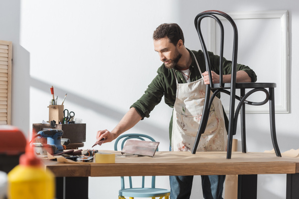 Bearded restorer in apron holding taking sandpaper near chair on table in workshop  - Foto, Bild