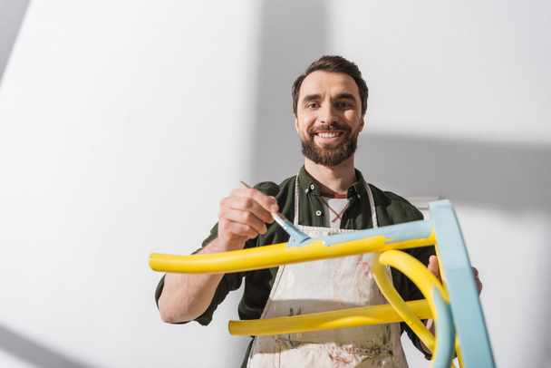 Smiling bearded restorer looking at camera while repainting chair in workshop  - Photo, Image