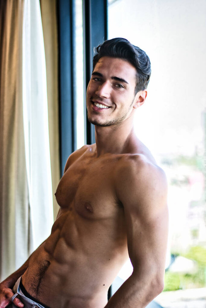 Sexy handsome young man standing shirtless in his bedroom next to window curtains - Foto, Bild