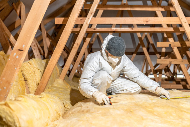 un hombre aísla el techo y el techo de la casa con lana de vidrio - Foto, Imagen