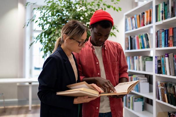 African American student shows information found in exam preparation book to project partner near bookshelves in college. Puzzled couple in university library looking concentrated busy in assignment. - Photo, Image