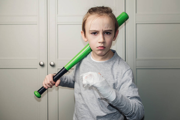 Boy with a broken arm in plaster holding baseball bat. Kid with broken hand after training. Sport time and healthcare. Health, lifestyle and sport concept. - Photo, Image