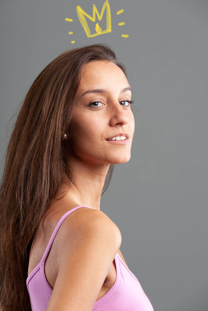 glance over her shoulder in a portrait of a slim young woman in a pink tank top and long hair with an expression of conceit and vanity augmented by a graphic crown on her head - concept of feeling sup - Foto, afbeelding