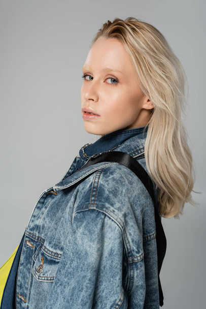young blonde model in stylish denim jacket looking at camera while posing isolated on grey  - Foto, immagini