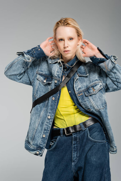 молодая блондинка в стильном джинсовом наряде смотрит в камеру, позируя с руками за головой, изолированными на сером  - Фото, изображение